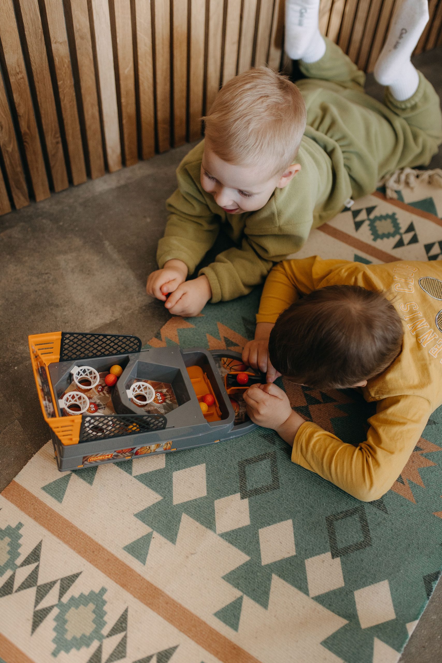 Two boys engaged in play with a toy indoors, lying on a patterned carpet.